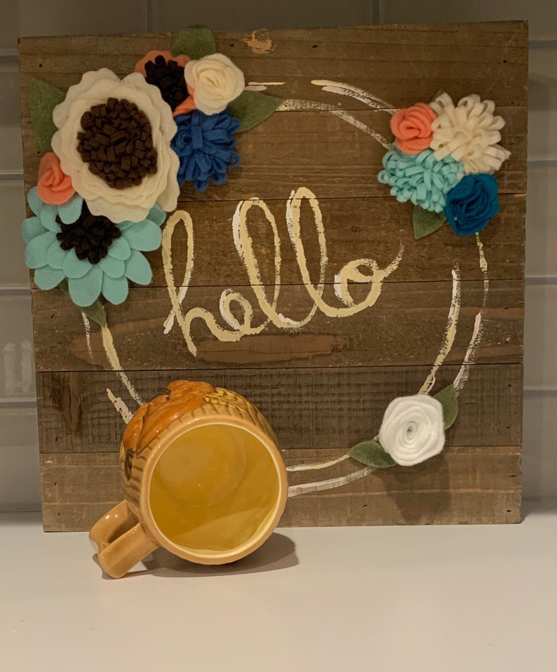 May include: A wooden sign with the word "hello" painted in yellow on it. The sign is decorated with a wreath of felt flowers in shades of white, blue, and pink. A yellow ceramic mug with a brown design is sitting in front of the sign.