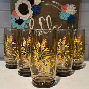 May include: Set of six vintage amber-tinted drinking glasses. Each glass features a gold wheat stalk design. A wooden sign with the word "hello" and felt flower accents is in the background.