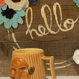 May include: A ceramic mug with a brown and yellow mushroom design. The mug has a handle and is sitting on a white surface. The background is a wooden surface with the word "hello" written in white paint.