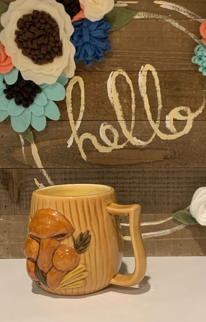 May include: A ceramic mug with a brown and yellow mushroom design. The mug has a handle and is sitting on a white surface. The background is a wooden surface with the word "hello" written in white paint.