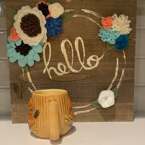 May include: A brown ceramic mug shaped like a tree trunk with a brown handle. The mug is in front of a wooden sign with the word "hello" written in white paint. The sign is decorated with felt flowers in shades of blue, white, and pink.