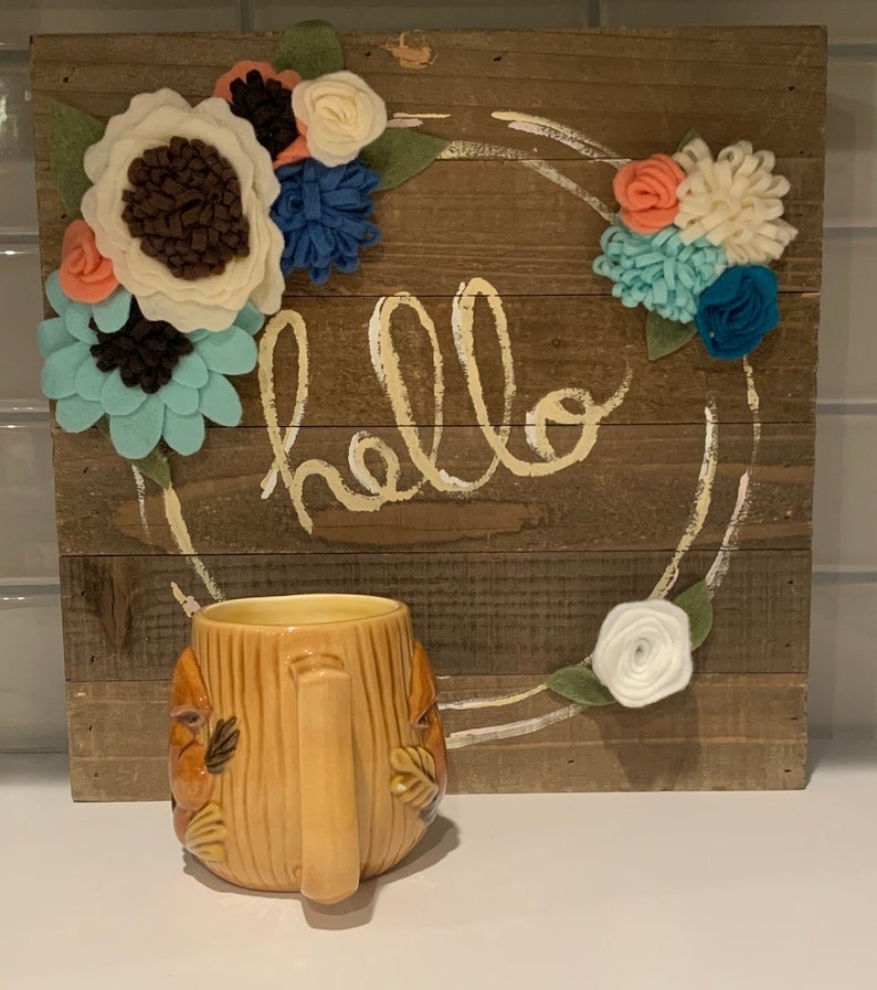 May include: A brown ceramic mug shaped like a tree trunk with a brown handle. The mug is in front of a wooden sign with the word "hello" written in white paint. The sign is decorated with felt flowers in shades of blue, white, and pink.