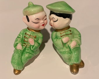 Könnte beinhalten: Zwei Keramikfiguren von zwei Personen in grüner und goldener Kleidung mit Hüten, die sich ansehen.