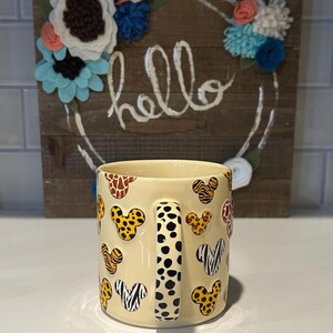 May include: A cream-colored ceramic mug featuring a pattern of Mickey Mouse heads with animal print designs. The handle has a black and white polka dot pattern. A wooden sign with the word "hello" and floral accents is in the background.