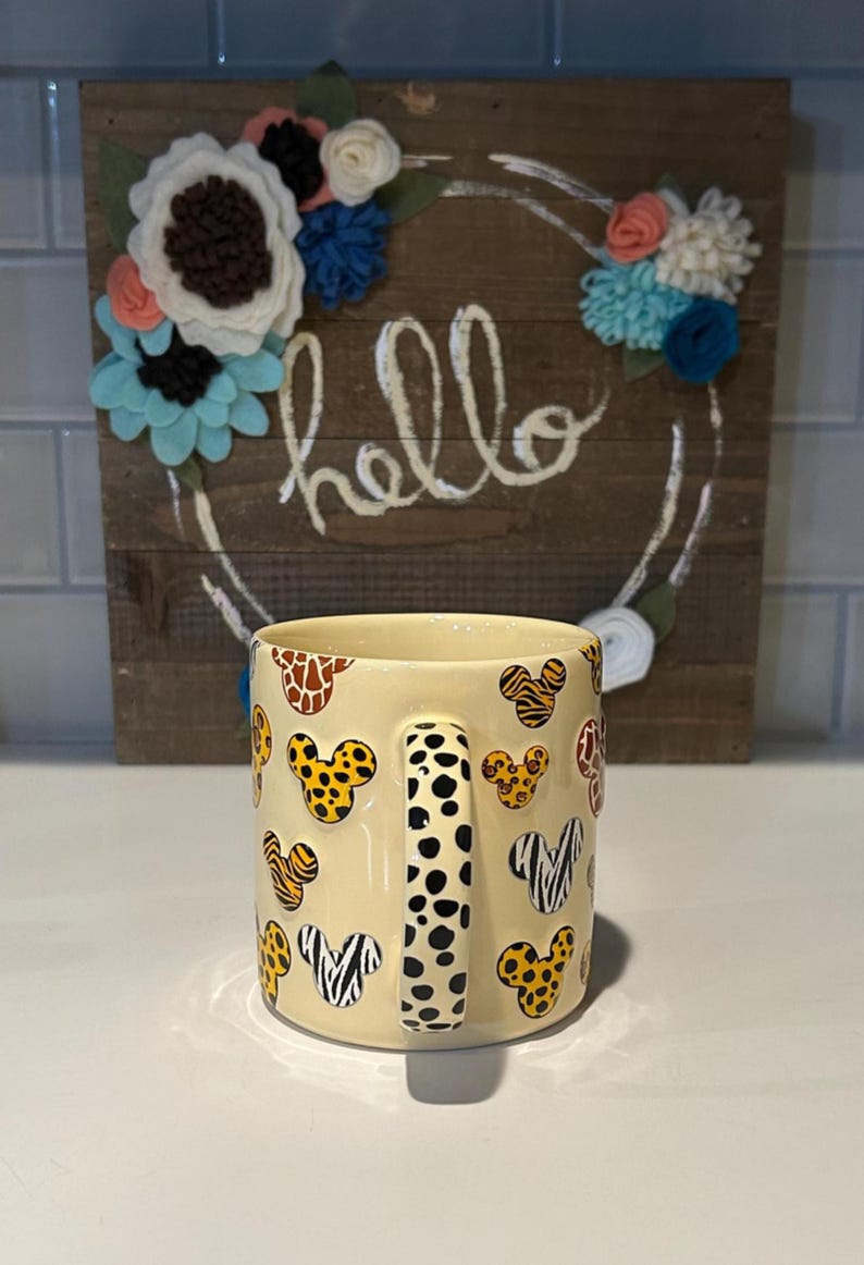 May include: A cream-colored ceramic mug featuring a pattern of Mickey Mouse heads with animal print designs. The handle has a black and white polka dot pattern. A wooden sign with the word "hello" and floral accents is in the background.