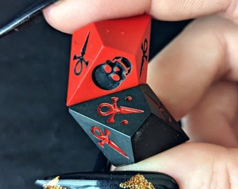 Puede incluir: Dos dados poliédricos, uno rojo y otro negro, son sostenidos. El dado rojo presenta una calavera, el negro un símbolo ankh. Ambos tienen detalles rojos. Estos dados son para juegos de mesa.