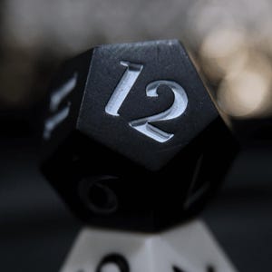 May include: Close-up of a black, multi-sided die with the number 12 in white. The die is resting on another die, with a blurred background. The die is likely used for tabletop games.