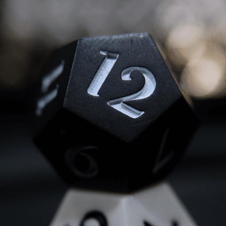 May include: Close-up of a black, multi-sided die with the number 12 in white. The die is resting on another die, with a blurred background. The die is likely used for tabletop games.