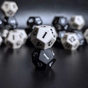 May include: Close-up of two twelve-sided dice, one white and one black, with the number one facing up on both. The white die is stacked on top of the black die. Other dice are blurred in the background.