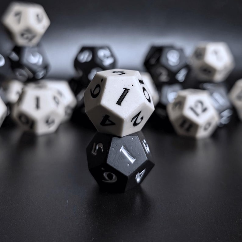 May include: Close-up of two twelve-sided dice, one white and one black, with the number one facing up on both. The white die is stacked on top of the black die. Other dice are blurred in the background.