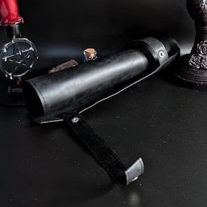 May include: A black leather cylindrical case with a strap and snap closure. The case is open, revealing the interior. A small glass bottle with a cork stopper is visible. The case is on a dark surface.