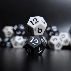May include: Two twelve-sided dice, one black and one white, stacked on top of each other. The top faces show the number 12. Other dice are in the background. These are used for tabletop role-playing games.