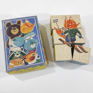 May include: A vintage wooden puzzle box with a colorful illustration of animals on the front. The box contains six wooden blocks with images of a fox skiing, a bear, a raccoon, a cat, a rabbit, and a blue bird.