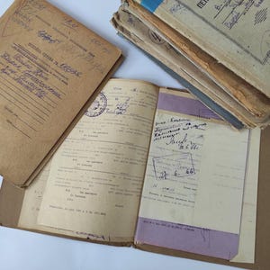 Può includere: Un documento vintage aperto con testo manoscritto e timbri, accanto a una pila di documenti simili. Il documento aperto mostra testo dettagliato e un timbro. I documenti sono in varie tonalità di marrone e beige, suggerendo l'età.
