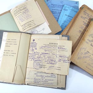 May include: A collection of vintage documents, including notebooks and loose papers, in various colors such as beige, blue, and green. The documents feature handwritten text, stamps, and signs of aging, suggesting historical significance. The items are arranged on a white surface.