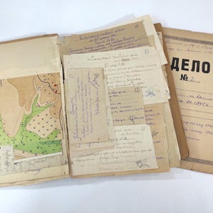 Puede incluir: Una carpeta de papel beige abierta que contiene varios documentos antiguos, notas manuscritas en cirílico y un mapa topográfico con secciones verdes y bronceadas. A la derecha se ve una cubierta beige con el texto en cirílico "ДЕЛО" y "№".