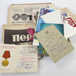 Könnte beinhalten: Eine Sammlung von Vintage-Dokumenten, Zeitungen und Karten. Die Artikel umfassen eine Zeitung mit kyrillischem Text, eine Urkunde mit einer Medaille und verschiedene Postkarten. Die Farbpalette wird von Beige, Rot und Schwarz dominiert.