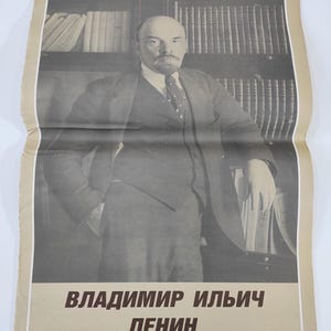 Può includere: Una fotografia in bianco e nero di Vladimir Il'ič Ul'janov, meglio conosciuto come Lenin, un rivoluzionario, politico e teorico politico russo. La foto mostra Lenin in piedi davanti a una libreria, con un abito e una cravatta. Il testo sotto la foto recita "Vladimir Il'ič Ul'janov, Vita e Attività."
