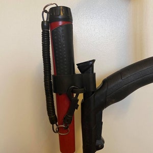 Op de afbeelding: Een rood-zwarte metaaldetector met een zwarte handgreep en een rode metalen schacht. De detector heeft een zwarte holster met een veerbelaste koord.