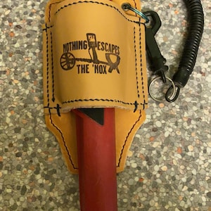 Puede incluir: Una funda de cuero marrón con un detector de metales rojo en su interior. La funda tiene una correa negra y un anillo de metal. La funda está bordada con el texto "NOTHING ESCAPES THE 'NOX'".