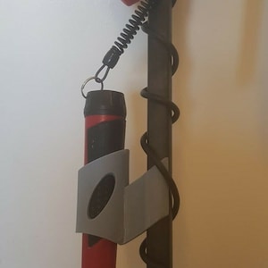 Op de afbeelding: Een rode en zwarte metaaldetector met een grijze kunststof houder voor een rode en zwarte metaaldetector sonde. De sonde is bevestigd aan de houder met een zwart snoer.