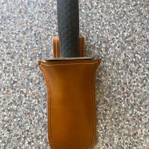 Könnte beinhalten: Eine braune Lederscheide mit einem genähten Rand, die für ein Gartengerät mit einem schwarzen Gummigriff ausgelegt ist.