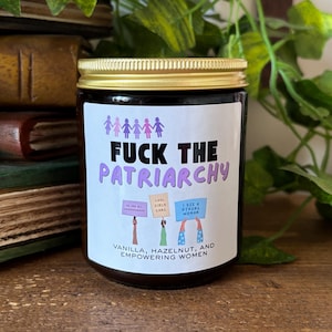 May include: A dark glass candle jar with a gold lid and a white label. The label features purple stick figures, the black and purple text FUCK THE PATRIARCHY, illustrations of people holding signs, and the text VANILLA, HAZELNUT, AND EMPOWERING WOMEN.