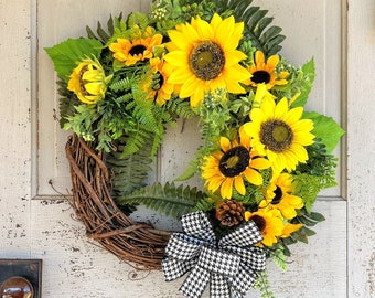 May include: A sunflower wreath with a black and white checkered bow. The wreath is made of grapevine and has a variety of greenery, including ferns and small leaves. The sunflowers are yellow with brown centers. The bow is tied around the wreath with a wire.