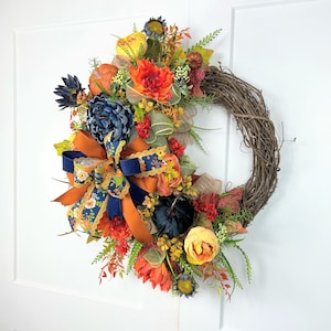 May include: A fall-themed wreath with a blue and orange ribbon, featuring faux pumpkins, sunflowers, and other autumnal foliage.