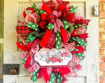 May include: A Christmas wreath with red and green plaid ribbon, red velvet ribbon, and a sign that says "Peace on Earth". The wreath also has a red truck with a white background.