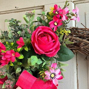 The hot pink cabbage roses are accented with pink wildflowers and beautiful greenery. The design cascades down the left side of the grapevine. The pink and burlap and white striped bow makes this everyday design look exquisite for any occasion.