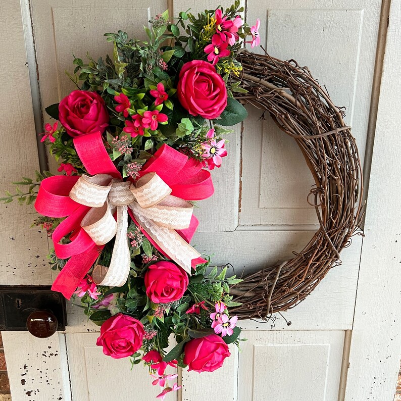 The hot pink cabbage roses are accented with pink wildflowers and beautiful greenery. The design cascades down the left side of the grapevine. The pink and burlap and white striped bow makes this everyday design look exquisite for any occasion.