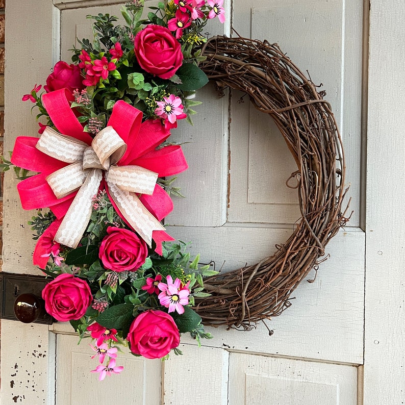 The hot pink cabbage roses are accented with pink wildflowers and beautiful greenery. The design cascades down the left side of the grapevine. The pink and burlap and white striped bow makes this everyday design look exquisite for any occasion.