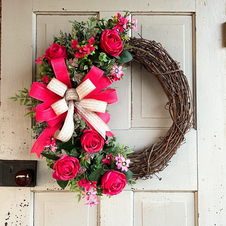 The hot pink cabbage roses are accented with pink wildflowers and beautiful greenery. The design cascades down the left side of the grapevine. The pink and burlap and white striped bow makes this everyday design look exquisite for any occasion.