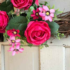 The hot pink cabbage roses are accented with pink wildflowers and beautiful greenery. The design cascades down the left side of the grapevine. The pink and burlap and white striped bow makes this everyday design look exquisite for any occasion.