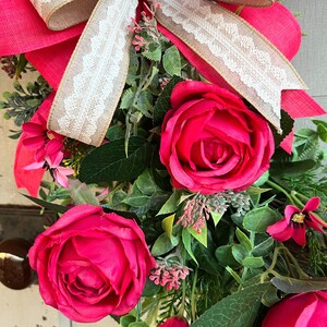 The hot pink cabbage roses are accented with pink wildflowers and beautiful greenery. The design cascades down the left side of the grapevine. The pink and burlap and white striped bow makes this everyday design look exquisite for any occasion.