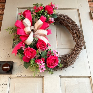 The hot pink cabbage roses are accented with pink wildflowers and beautiful greenery. The design cascades down the left side of the grapevine. The pink and burlap and white striped bow makes this everyday design look exquisite for any occasion.