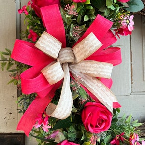 The hot pink cabbage roses are accented with pink wildflowers and beautiful greenery. The design cascades down the left side of the grapevine. The pink and burlap and white striped bow makes this everyday design look exquisite for any occasion.