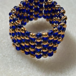 K&ouml;nnte beinhalten: Ein handgefertigter Ring aus abwechselnd kleinen, runden, goldenen und blauen Perlen. Der Ring besteht aus mehreren Reihen von Perlen, wodurch ein strukturiertes, bandartiges Aussehen entsteht. Der Ring wird auf einer wei&szlig;en Oberfl&auml;che gezeigt.