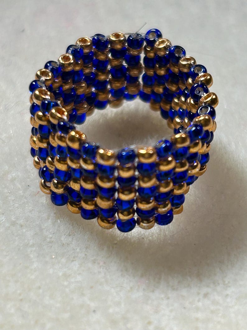 K&ouml;nnte beinhalten: Ein handgefertigter Ring aus abwechselnd kleinen, runden, goldenen und blauen Perlen. Der Ring besteht aus mehreren Reihen von Perlen, wodurch ein strukturiertes, bandartiges Aussehen entsteht. Der Ring wird auf einer wei&szlig;en Oberfl&auml;che gezeigt.