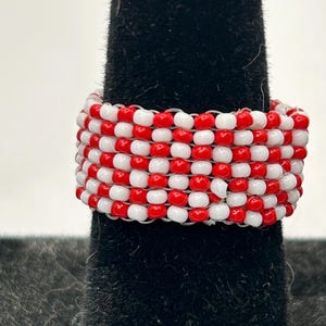 K&ouml;nnte beinhalten: Ein handgefertigter Ring mit abwechselnd roten und wei&szlig;en Perlen. Der Ring wird auf einem schwarzen Samtuntergrund pr&auml;sentiert, der die lebendigen Farben und die aufwendige Perlenarbeit hervorhebt. Ein einzigartiges Schmuckst&uuml;ck.