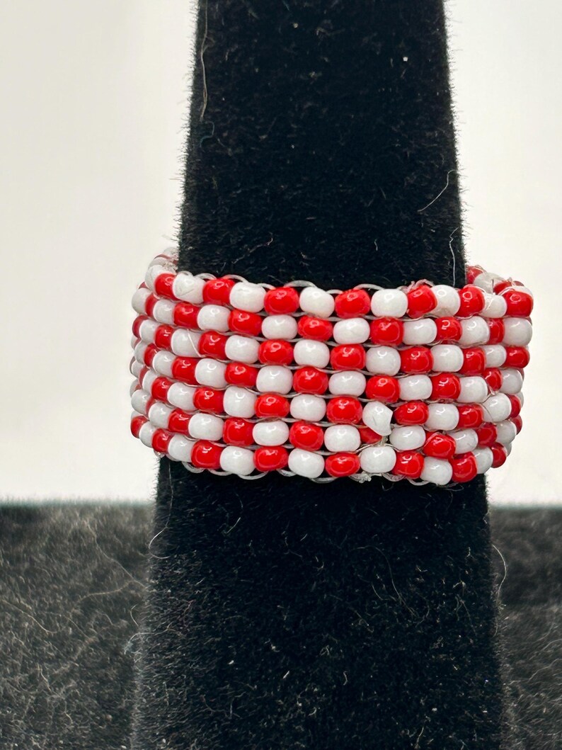 K&ouml;nnte beinhalten: Ein handgefertigter Ring mit abwechselnd roten und wei&szlig;en Perlen. Der Ring wird auf einem schwarzen Samtuntergrund pr&auml;sentiert, der die lebendigen Farben und die aufwendige Perlenarbeit hervorhebt. Ein einzigartiges Schmuckst&uuml;ck.