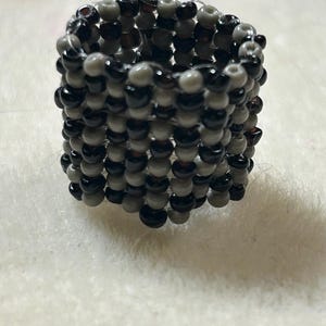 K&ouml;nnte beinhalten: Ein handgefertigter Ring aus kleinen, runden Perlen in den Farben Schwarz, Grau und Dunkelgrau. Der Ring hat eine zylindrische Form und liegt auf einer wei&szlig;en, strukturierten Oberfl&auml;che.