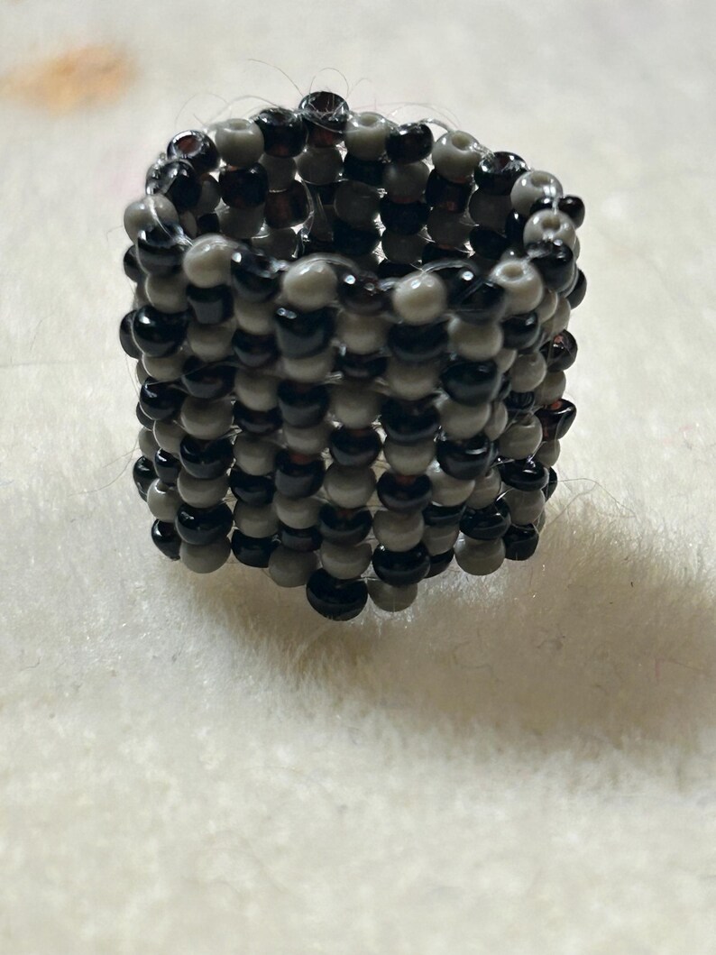 K&ouml;nnte beinhalten: Ein handgefertigter Ring aus kleinen, runden Perlen in den Farben Schwarz, Grau und Dunkelgrau. Der Ring hat eine zylindrische Form und liegt auf einer wei&szlig;en, strukturierten Oberfl&auml;che.