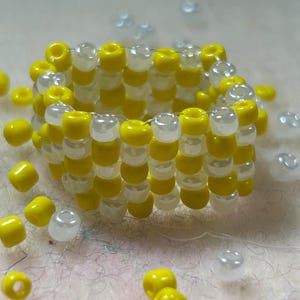 K&ouml;nnte beinhalten: Ein handgefertigter Perlenring mit abwechselnden Reihen aus leuchtend gelben und durchscheinend wei&szlig;en Perlen. Der Ring ist kreisf&ouml;rmig, zylindrisch geformt und von losen Perlen in den gleichen Farben umgeben. Die Perlen sind klein und gleichm&auml;&szlig;ig angeordnet.