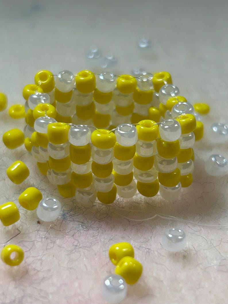 K&ouml;nnte beinhalten: Ein handgefertigter Perlenring mit abwechselnden Reihen aus leuchtend gelben und durchscheinend wei&szlig;en Perlen. Der Ring ist kreisf&ouml;rmig, zylindrisch geformt und von losen Perlen in den gleichen Farben umgeben. Die Perlen sind klein und gleichm&auml;&szlig;ig angeordnet.