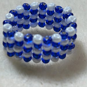 K&ouml;nnte beinhalten: Ein Perlenring mit abwechselnden Reihen aus wei&szlig;en und blauen Perlen. Der Ring besteht aus kleinen, runden Perlen, wodurch eine strukturierte Oberfl&auml;che entsteht. Die Farbkombination bildet einen auff&auml;lligen Kontrast, wobei die blauen Perlen gl&auml;nzend wirken.