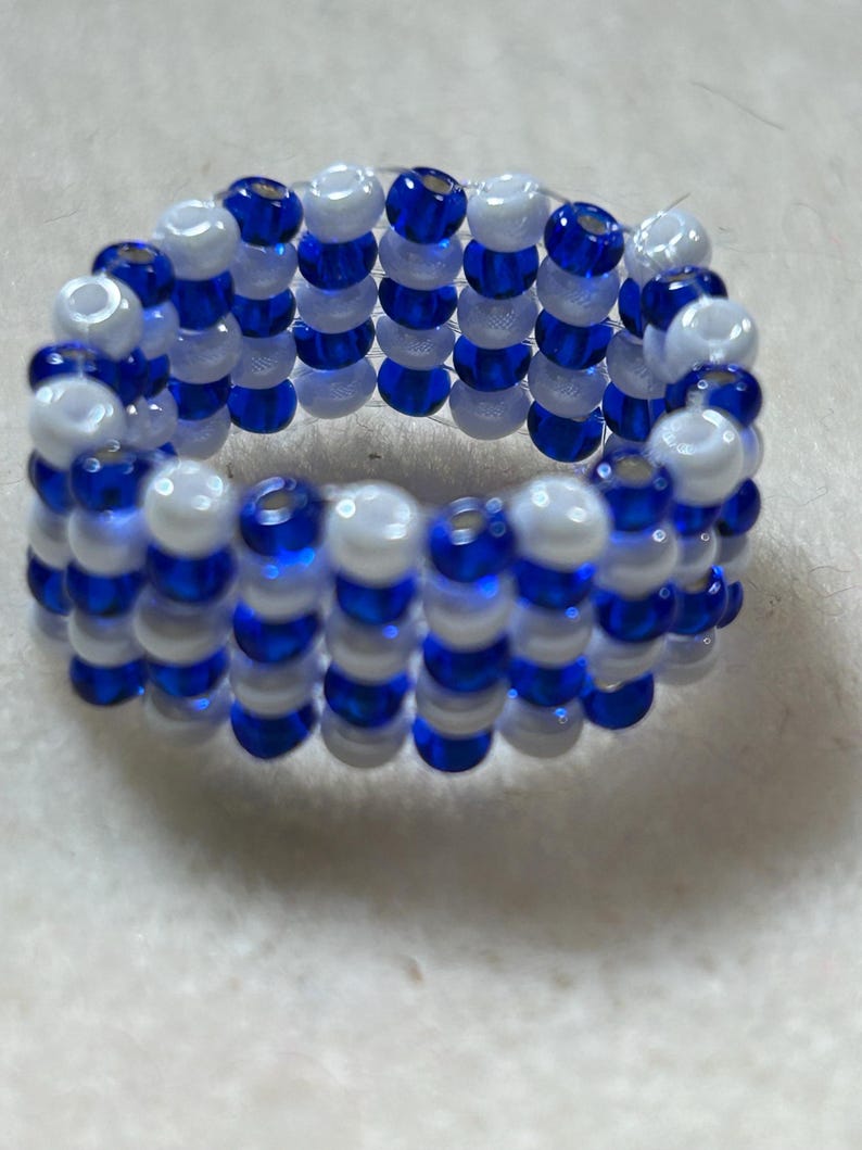 K&ouml;nnte beinhalten: Ein Perlenring mit abwechselnden Reihen aus wei&szlig;en und blauen Perlen. Der Ring besteht aus kleinen, runden Perlen, wodurch eine strukturierte Oberfl&auml;che entsteht. Die Farbkombination bildet einen auff&auml;lligen Kontrast, wobei die blauen Perlen gl&auml;nzend wirken.