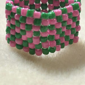 K&ouml;nnte beinhalten: Ein handgefertigter Ring aus rosa und gr&uuml;nen Perlen. Die Perlen sind in einem Schachbrettmuster miteinander verwoben, wodurch ein strukturiertes, farbenfrohes Accessoire entsteht. Der Ring ist ein lebendiges Schmuckst&uuml;ck.