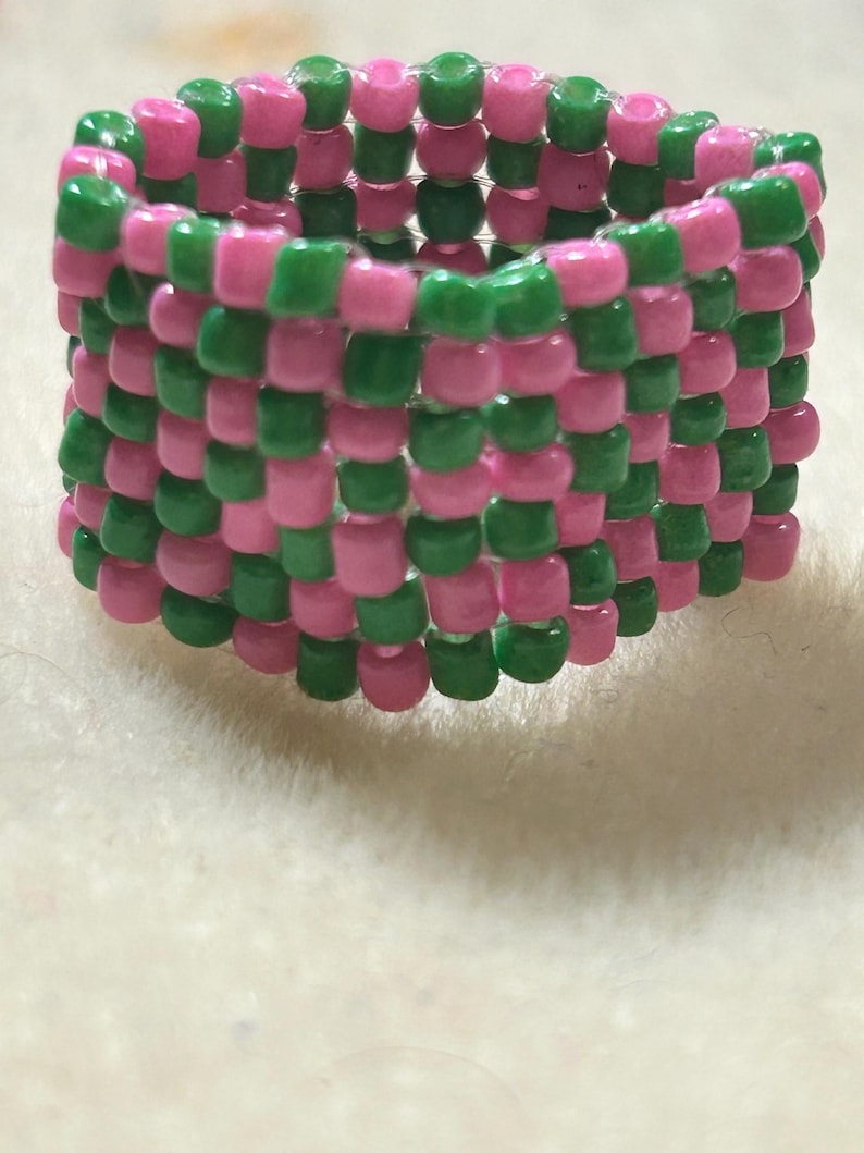 K&ouml;nnte beinhalten: Ein handgefertigter Ring aus rosa und gr&uuml;nen Perlen. Die Perlen sind in einem Schachbrettmuster miteinander verwoben, wodurch ein strukturiertes, farbenfrohes Accessoire entsteht. Der Ring ist ein lebendiges Schmuckst&uuml;ck.